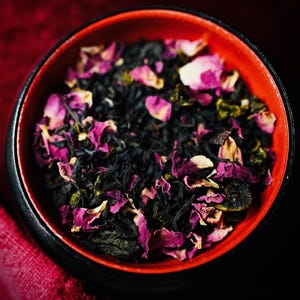 May include: A small round bowl with a black exterior and a vibrant red interior filled with a blend of loose leaf black tea, dried green tea leaves, and scattered bright pink and white rose petals, creating a colourful and textured botanical mixture.