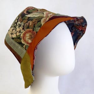 May include: A bonnet featuring a multi-colored floral tapestry crown with a dark orange velvet brim and a sage green velvet side panel. The interior lining displays a dark blue fabric with a repeating floral pattern, creating a layered look.
