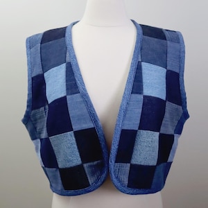 May include: A denim vest with a patchwork design in various shades of blue. The vest is sleeveless and has a V-neck. The edges are trimmed with a darker blue fabric. The vest is displayed on a white mannequin.
