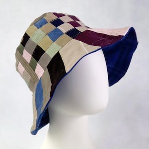 May include: A patchwork bucket hat with a wide brim, displayed on a white mannequin head. The hat features a variety of colors including blue, pink, beige, and purple, arranged in a checkered pattern. The brim's underside is a vibrant blue.