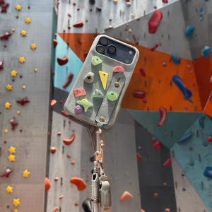 May include: A light gray phone case featuring small, colorful climbing hold decorations and a vertical strap with metallic rivets. The case is attached to a silver metal carabiner and a lanyard, designed to resemble a rock climbing wall surface.