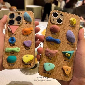 May include: Two cork-textured phone cases featuring various colorful, three-dimensional rock climbing holds attached to the back. Each hold is labeled with terms like Side pull, Volume, Jug, Pocket, Crimp, Pinch, Sloper, and Undercling in black ink.