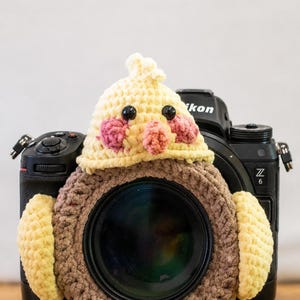Op de afbeelding: Een zwarte Nikon Z 6 camera met een gehaakte vogel lens cover. De vogel cover is geel, bruin en roze, met zwarte knoopogen. De lens cover is gevormd als een vogel met vleugels en een kuif.