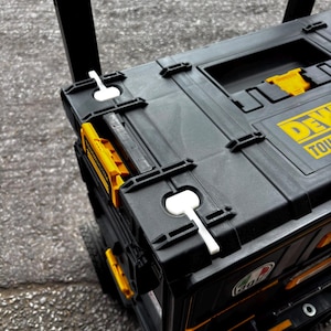 Può includere: Una cassetta degli attrezzi mobile DeWalt ToughSystem nera e gialla con struttura in plastica resistente, chiusure laterali integrate, una maniglia superiore e due linguette di bloccaggio bianche sulla superficie superiore.
