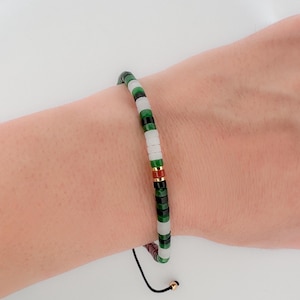 May include: A beaded bracelet featuring a mix of green, white, and gold beads. The bracelet is strung on a black cord with a gold-colored bead at the end. The beads are small and round, creating a minimalist design.