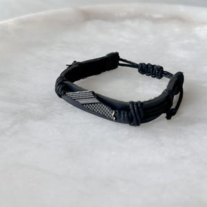 May include: Black leather bracelet with a woven silver-tone metal accent. The bracelet features a braided cord closure and is displayed on a white surface. This accessory is suitable for everyday wear.