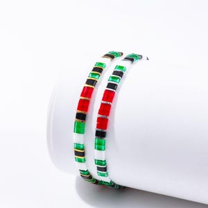 May include: Two beaded bracelets featuring a repeating pattern of red, black, green, white, and gold beads. The bracelets are curved and displayed on a white cylindrical surface, highlighting the colourful design.