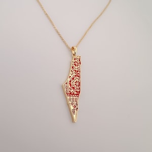 May include: A gold-colored necklace with a pendant in the shape of a country map, filled with red and white patterned designs. The necklace hangs against a white background. The brand name "Alluria Jewelry" is at the bottom.