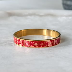 May include: A gold-toned bangle bracelet with a red geometric pattern. The bracelet has a smooth, polished finish and a classic design. The red pattern is a repeating design of squares and floral shapes.