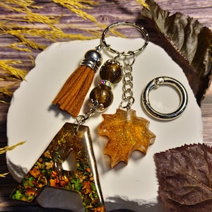 Può includere: Un portachiavi con una lettera A in resina riempita di foglie autunnali essiccate, un ciondolo a forma di foglia d'acero in resina glitterata dorata, due perline marrone scuro e una nappa in camoscio marrone su un anello in metallo.