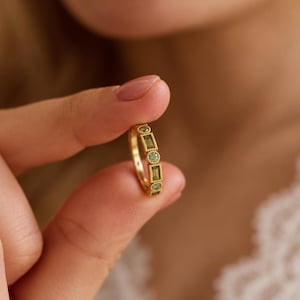 May include: A gold ring featuring an alternating pattern of small round green stones and rectangular green baguette stones. The metal band has a beaded texture surrounding each stone, creating a detailed and structured design on the ring's surface.