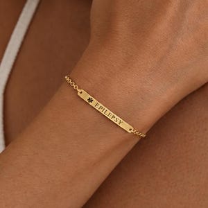 Könnte beinhalten: Ein goldfarbenes medizinisches Notfallarmband mit einem dünnen, rechteckigen Metallsteg, der mit dem Wort EPILEPSY und einem medizinischen Stern-Symbol graviert ist. Der Steg ist an einer feinen goldfarbenen Kette befestigt.