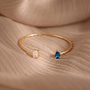 Puede incluir: Una pulsera abierta de tono dorado con dos engastes de piedras preciosas distintos en los extremos. Un extremo sostiene una piedra transparente de corte esmeralda rectangular, mientras que el otro presenta una piedra azul vibrante en forma de pera.