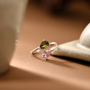 May include: A silver-toned open ring featuring two pear-cut gemstones. One stone is a deep olive green, and the other is a soft pastel pink. The band is slender and metallic, designed with an adjustable gap between the two teardrop-shaped stones.
