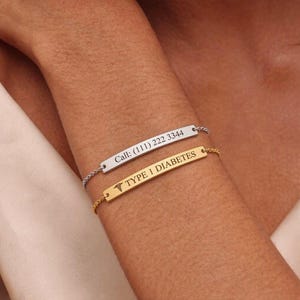 Pode incluir: Duas pulseiras de barra metálica com correntes finas. A barra prateada está gravada com o texto Call: (111) 222 3344. A barra dourada apresenta um símbolo médico de caduceu e o texto gravado TYPE 1 DIABETES. Ambas são usadas juntas.