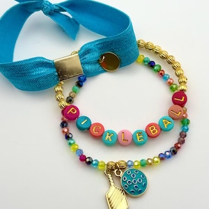 May include: A set of two bracelets featuring a bright blue elastic hair tie with a gold square accent and a beaded bracelet. The beaded bracelet includes gold-toned spacers, multicolored faceted beads, and round beads spelling PICKLEBALL.