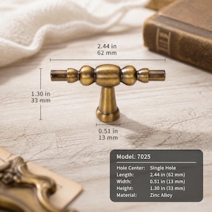 May include: A T-shaped cabinet knob in an antique brass finish with a central post and rounded decorative accents. The knob measures 2.44 inches in length, 1.30 inches in height, and 0.51 inches in width. The text indicates Model 7025, single hole.
