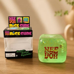 Puede incluir: Un cubo blando de color verde lima translúcido con purpurina multicolor incrustada en su interior. La palabra NEE DOH está impresa en rojo y amarillo en la parte frontal. Junto a él hay una caja de cartón negra etiquetada NEE DOH NICE CUBE.