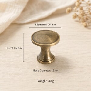 Pode incluir: Um puxador de móvel redondo com acabamento em latão antigo e uma superfície superior lisa e ligeiramente côncava. O puxador tem um diâmetro de 25 mm, uma altura de 25 mm e um diâmetro de base de 15 mm. O peso total da peça é de 30 gramas.