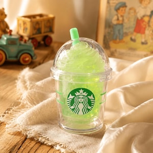May include: A clear plastic tumbler with a dome lid and a light green straw, filled with a bright green textured substance resembling a frozen beverage. The cup features a green circular Starbucks logo on the front and two horizontal green lines.