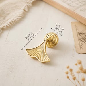 Pode incluir: Um puxador de móvel em metal dourado com formato de folha de ginkgo e base circular. O puxador mede 52 mm de comprimento e 24 mm de largura, conforme indicado pelas linhas de medição e etiquetas de texto visíveis na imagem.