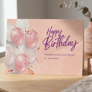 Może przedstawiać: Prostokątna kartka okolicznościowa z delikatnym brzoskwiniowym tłem i grupą różowych oraz białych balonów. Tekst głosi Happy Birthday, I remembered the card. The gift is... somewhere. Kartka jest wyeksponowana na drewnianej powierzchni.