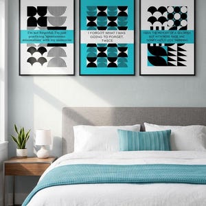 May include: Three framed art prints with black frames. Each print features a geometric design in black and white or teal, with text in a contrasting color. The prints are displayed above a bed with white linens and a teal throw.