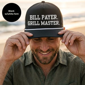 May include: A black baseball cap with a white rope detail across the base of the crown. The front of the cap features the white text "BILL PAYER. GRILL MASTER." in a bold, sans-serif font. The cap has a structured five-panel design.