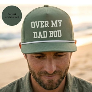 May include: An olive green five-panel baseball cap featuring a white rope detail across the base of the crown and the text OVER MY DAD BOD printed in bold white capital letters on the front panel. The cap has a structured front and flat brim.