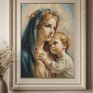 Op de afbeelding: Een ingelijst kruissteekborduurwerk met een vrouw met lang blond haar, een blauwe hoofdbedekking en een bloemenkrans, die een jong kind vasthoudt. Het kunstwerk heeft zachte pasteltinten en zit in een sierlijke, lichtgekleurde lijst.