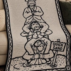 May include: A black and white crocheted blanket featuring a graphic design of three people stacked vertically, with the top person wearing a hat. A sign to the right of the figures reads QUICK SAND. The blanket has a decorative geometric border.