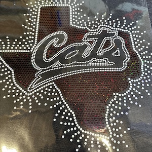 May include: A heat transfer decal featuring a rhinestone design of the state of Texas with a sunburst pattern around the border. The word Cats is written in cursive script across the center in white rhinestones against a dark red glitter background.