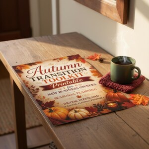 May include: A rectangular printable document titled Autumn TRANSITION TOOLKIT Printable for NEW BUSINESS OWNERS, featuring illustrations of pumpkins and autumn leaves, placed next to a green mug on a dark red knitted coaster.