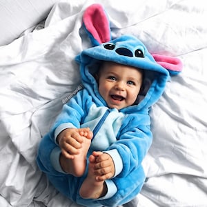 May include: A soft, light blue plush onesie featuring a hood with large, upright pink-lined ears and a character face with black eyes and a nose. The garment has long sleeves with light blue cuffs and a central zipper closure, designed for comfort.