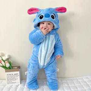May include: A plush, light blue one-piece jumpsuit featuring a hood with large, upright ears that have bright pink inner linings. The jumpsuit has a central zipper closure, a lighter blue belly panel, and embroidered eyes and nose on the hood.