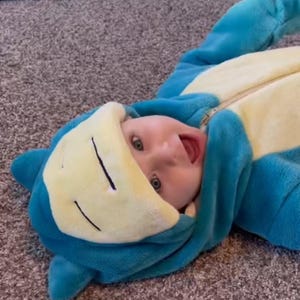 May include: A plush, soft-textured onesie featuring a character design with a blue body and a cream-colored belly. The hood includes a character face with closed eyes and ears, while the front of the garment has a central zipper closure.