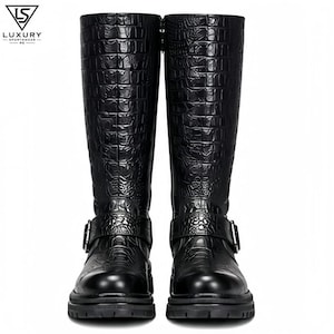 May include: A pair of black knee-high boots featuring a glossy crocodile-embossed texture, a rounded toe, and a buckled strap across the ankle. The boots include a thick lug sole and a side zip. The logo LS LUXURY SPORTSWEAR PK is visible.
