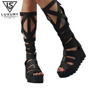 Puede incluir: Sandalias romanas negras hasta la rodilla con suela de plataforma gruesa y dentada, diseño de punta abierta y una serie de tiras anchas entrecruzadas que se extienden desde el pie hasta la pantorrilla, con el texto LUXURY SPORTSWEAR PK.