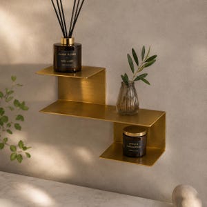 Könnte beinhalten: Ein goldfarbenes Wandregal aus Metall mit abgestuftem Design. Auf der obersten Ebene steht ein dunkelbrauner Zara Home Dark Amber Diffusor. Die mittlere Ebene enthält eine klare Glasvase mit grünen Blättern. Die untere Ebene trägt eine Cedar & Sandalwood Kerze.