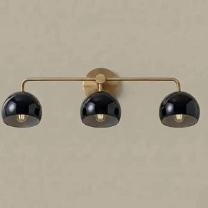 May include: A three-light wall fitting featuring a horizontal brass arm and a circular wall mount. The fitting includes three glossy black dome-shaped shades, each housing a single light bulb, arranged in a linear configuration.