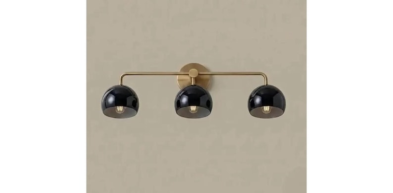May include: A three-light wall fitting featuring a horizontal brass arm and a circular wall mount. The fitting includes three glossy black dome-shaped shades, each housing a single light bulb, arranged in a linear configuration.