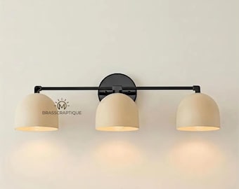 May include: A three-light wall sconce featuring a horizontal black metal bar and a circular black wall mount. It includes three dome-shaped beige shades directed downward, each housing a light bulb. The text BRASSCRAPTIQUE is visible.
