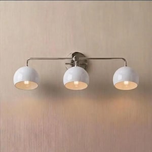 May include: A three-light wall fitting featuring a horizontal brushed nickel metal bar and a circular wall mount. Three white dome-shaped glass shades are attached to the bar, each housing a single light bulb that emits a warm glow.