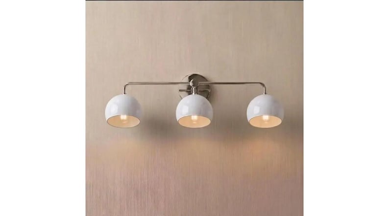 May include: A three-light wall fitting featuring a horizontal brushed nickel metal bar and a circular wall mount. Three white dome-shaped glass shades are attached to the bar, each housing a single light bulb that emits a warm glow.
