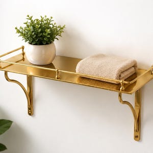 May include: A gold-colored metal wall shelf with a polished finish, featuring a protective railing along the edges and two curved support brackets. A small white ceramic pot containing a green plant and a folded beige towel sit on the shelf.