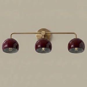 May include: A wall-mounted light fitting featuring a horizontal brass arm and a circular backplate. Three dome-shaped shades in a deep burgundy finish are attached to the arm, each housing a single light bulb directed downwards.
