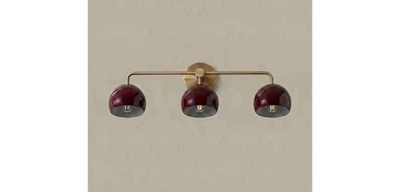 May include: A wall-mounted light fitting featuring a horizontal brass arm and a circular backplate. Three dome-shaped shades in a deep burgundy finish are attached to the arm, each housing a single light bulb directed downwards.