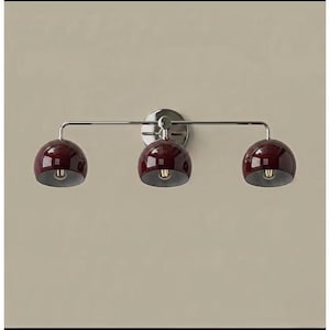 May include: A wall-mounted light fitting featuring three glossy, deep burgundy, dome-shaped shades attached to a polished chrome horizontal bar. The fitting includes a circular chrome wall plate at the centre, supporting the three individual light heads.