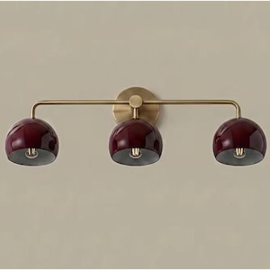 May include: A wall-mounted light fitting featuring a horizontal brass bar with three downward-facing, glossy deep burgundy dome-shaped shades. The fitting is attached to the wall by a circular brass backplate centred behind the middle light.