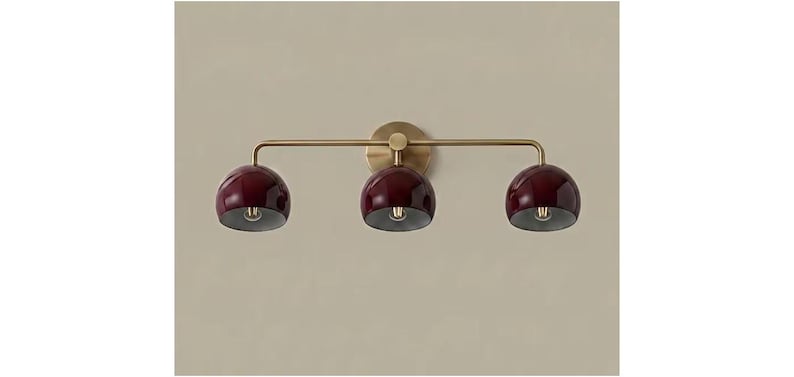 May include: A wall-mounted light fitting featuring a horizontal brass bar with three downward-facing, glossy deep burgundy dome-shaped shades. The fitting is attached to the wall by a circular brass backplate centred behind the middle light.